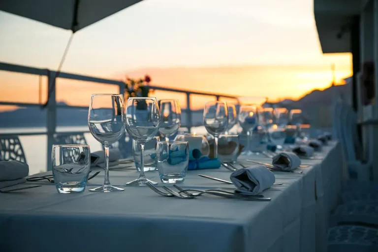 Elegant outdoor dining setup at dusk in Ibiza, featuring a line of neatly set tables with clear wine glasses, overlooking a serene view of the ocean and mountains under a soft sunset.