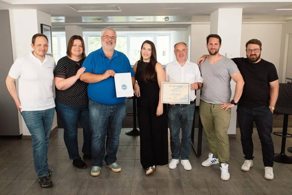 Seven Avcon Jet Austria team members—Bernhard Seedoch, Alicja Froböse, Krisztian Szekacs, Alexandra Stefa, Christian Hrauda, Marcus Degasperi, and Markus Grafl— celebrating the renewal of their Wyvern Wingman and IS-BAO Stage 2 certifications. Krisztian Szekacs holds a certificate labeled “IS-BAO,” and Christian Hrauda displays a framed Wyvern certificate. Smiling and standing in casual attire, the group marks a milestone for Avcon Jet Austria's ongoing commitment to aviation safety and quality.