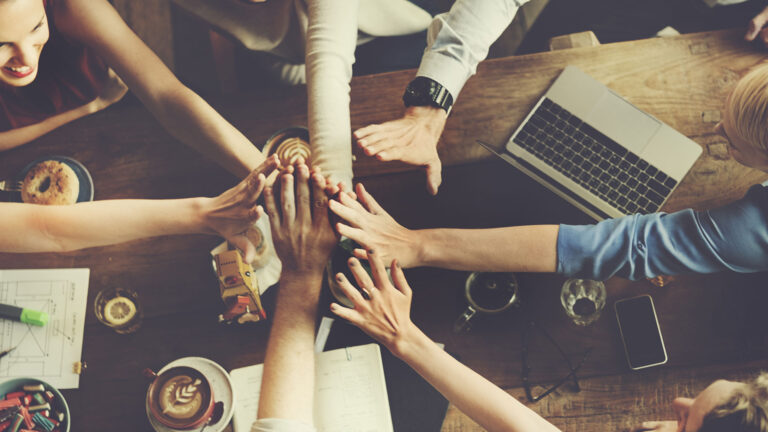 Team of employees high-fiving over a desk with different office materials, coffee and a notebook